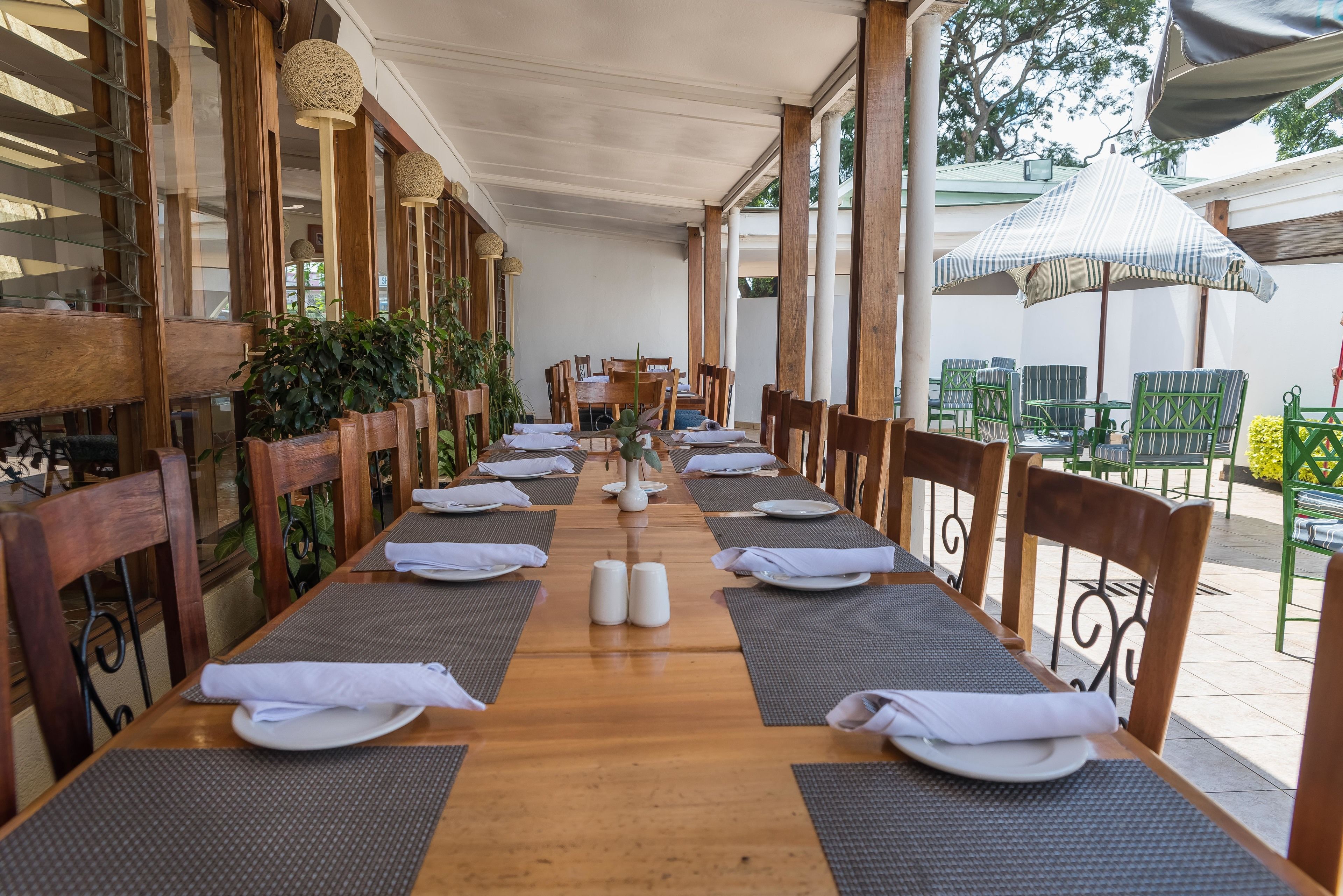 A long wooden table set with white napkins, plates, and candles, surrounded by chairs and plants, under a covered patio area with umbrellas.
