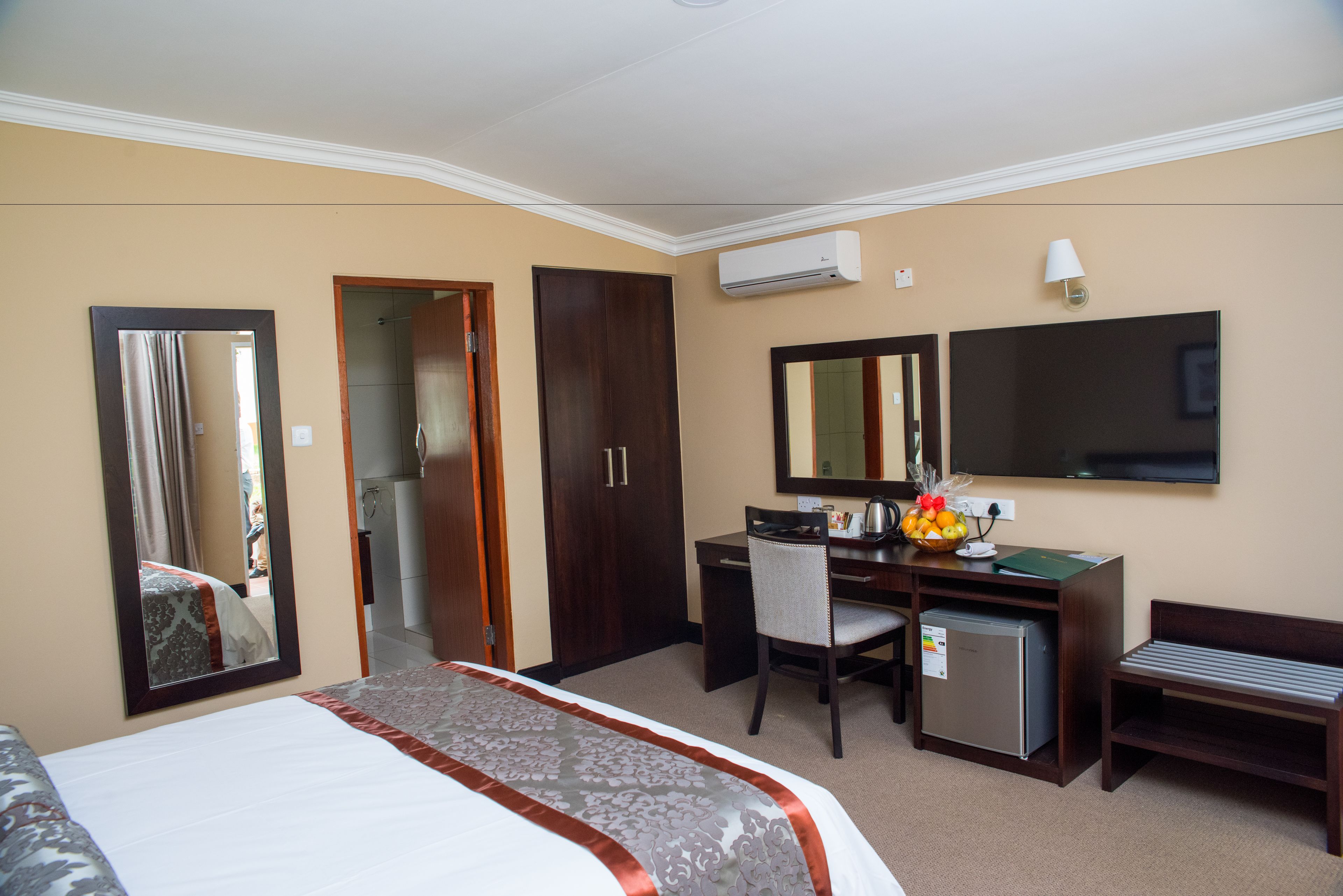 A hotel room with a large bed, a desk with a computer, a small refrigerator, a bench, a mirror, and a television.