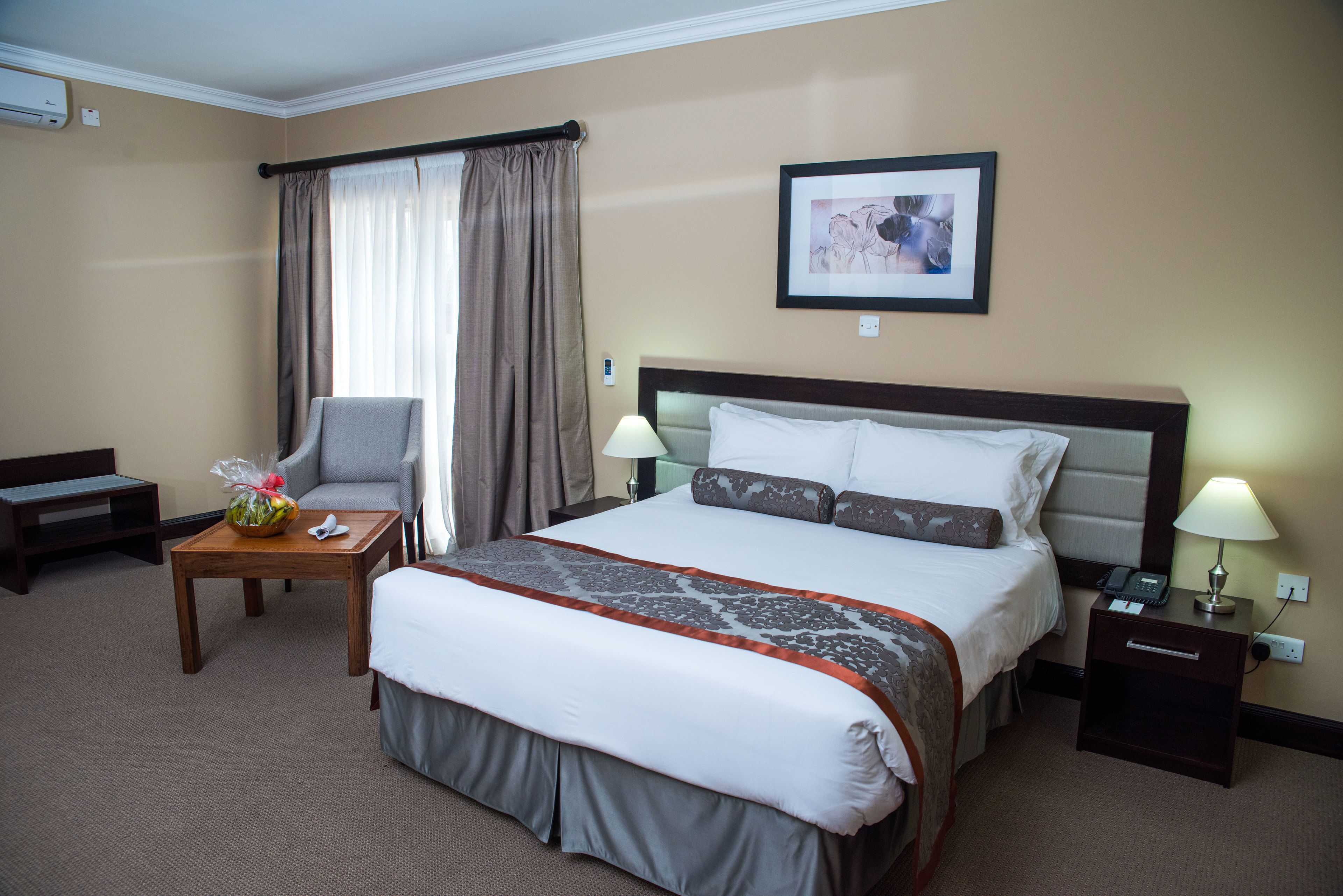 A hotel room with a large bed, a bedside table with a lamp, a chair, a small table with a fruit bowl, and a framed picture on the wall.
