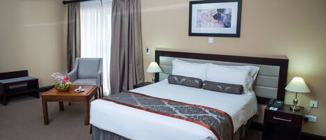 A hotel room with a large bed, a bedside table with a lamp, a chair, a small table with a fruit bowl, and a framed picture on the wall.