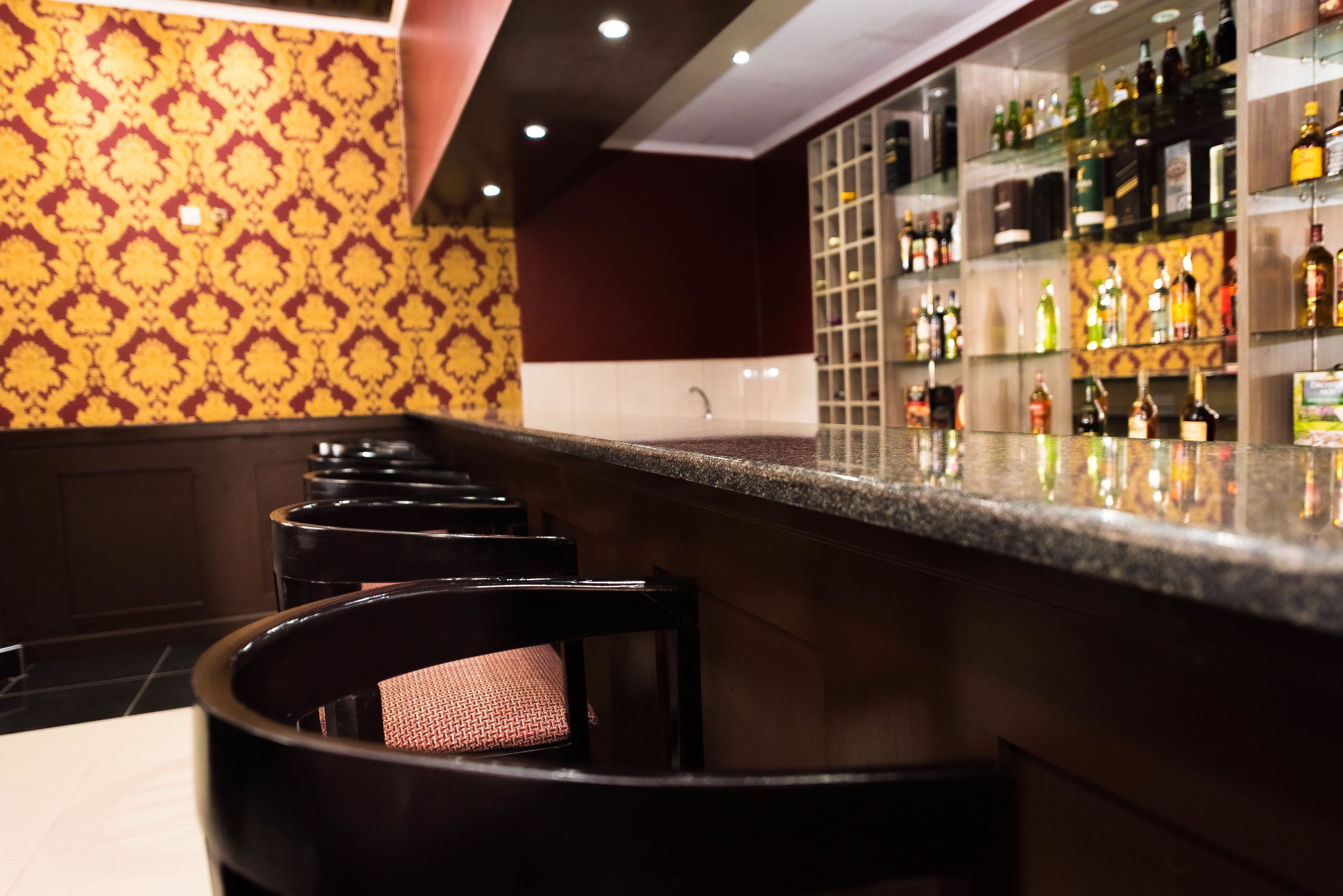 A bar area with a patterned wall, a counter with bottles, and bar stools.