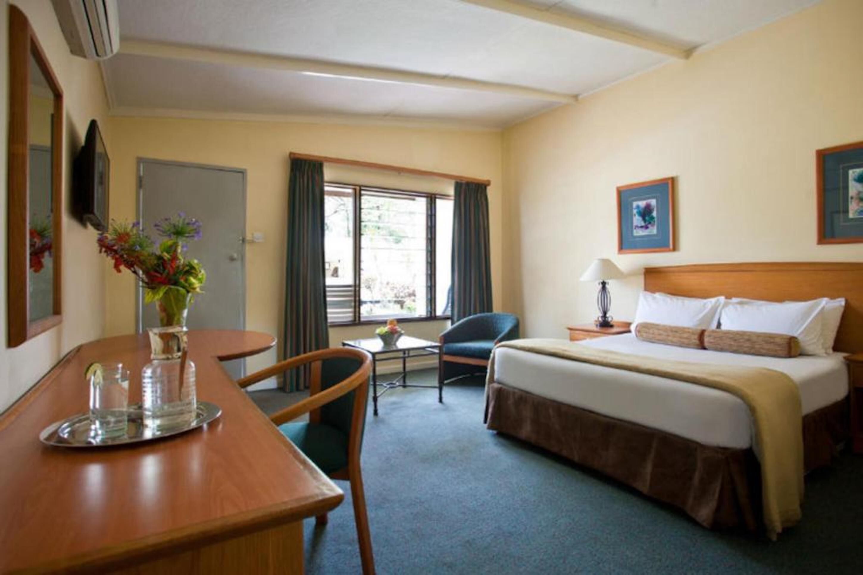 A hotel room with a bed, a desk with a chair, a small table, a vase with flowers, and a window with curtains.