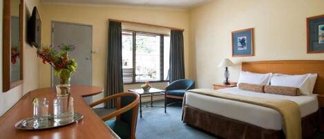A hotel room with a bed, a desk with a chair, a small table, a vase with flowers, and a window with curtains.