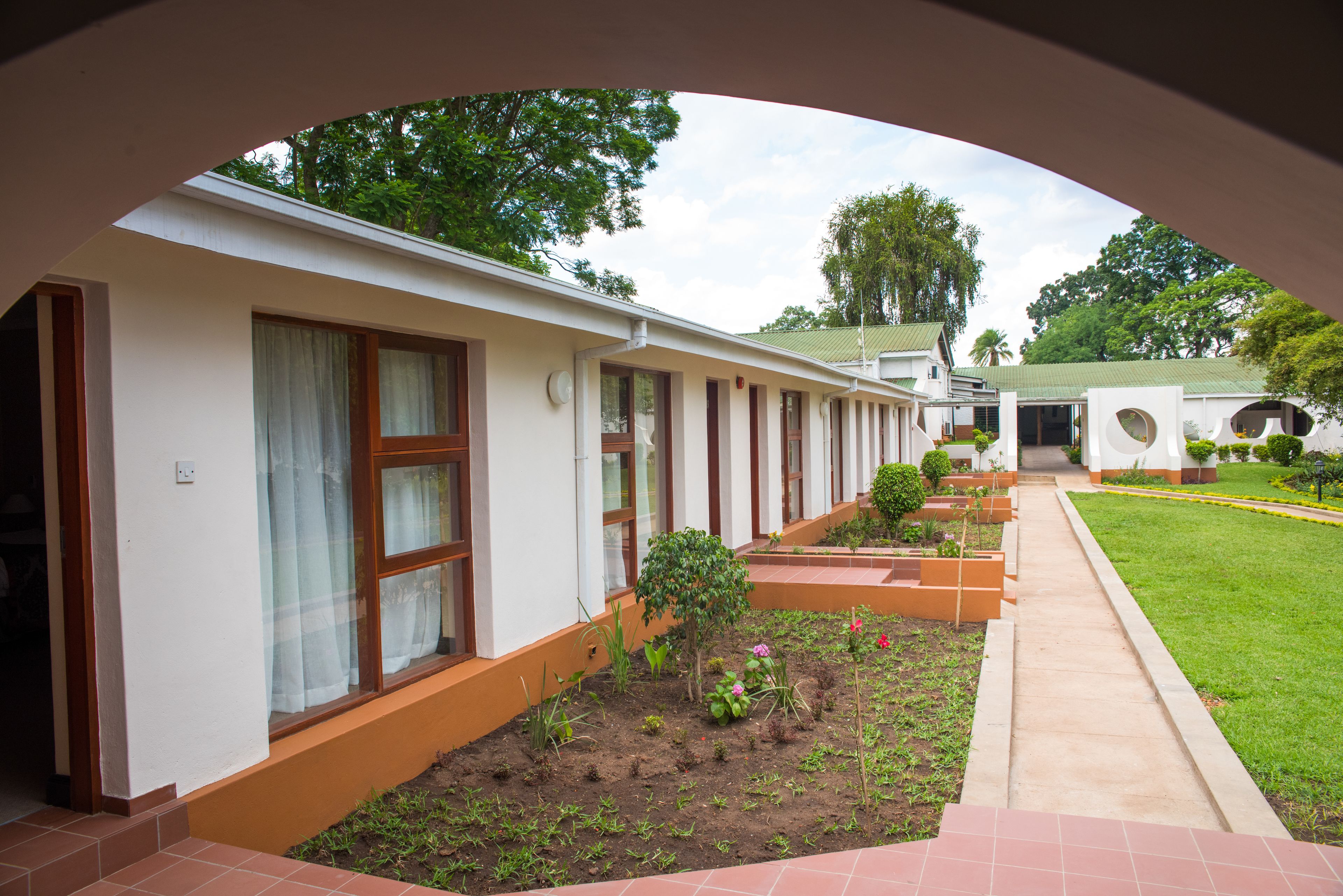 A corridor with a row of rooms, each with a window and a door. There is a garden area with plants and a pathway leading to a building entrance.