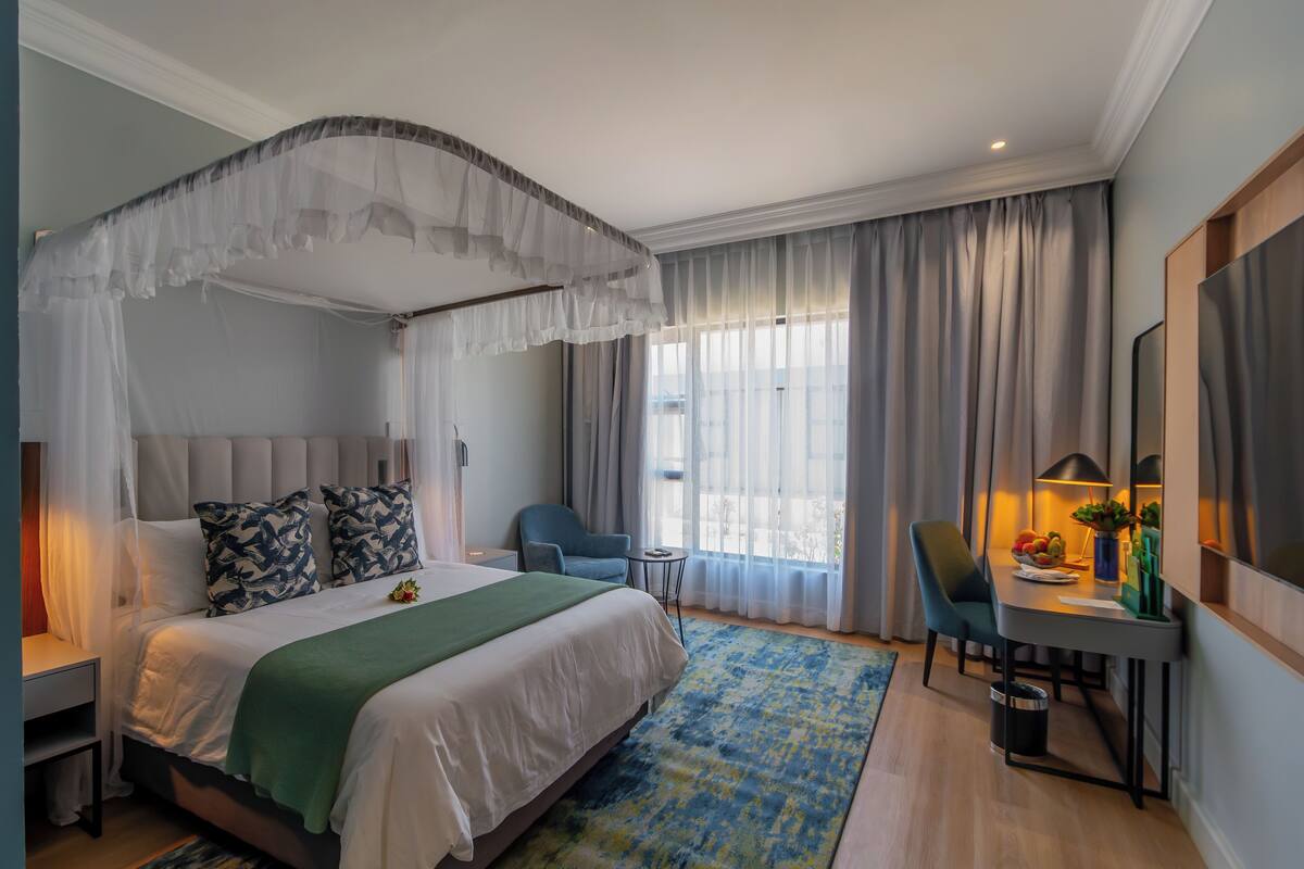 A hotel room with a bed, bedside table, chair, desk with a lamp, and a large window with curtains.
