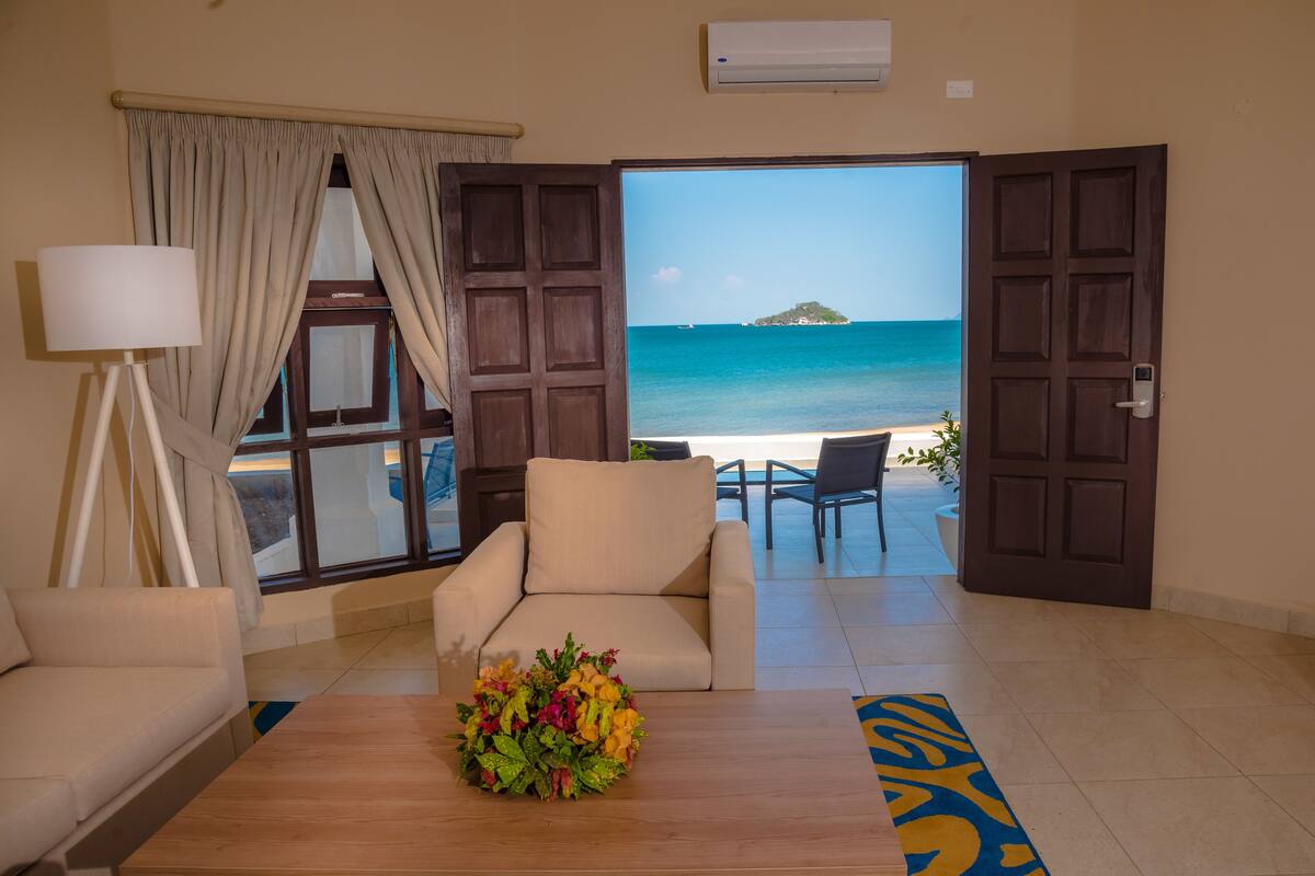A hotel room with a view of the beach, a sofa, a table with a flower arrangement, and a lamp.