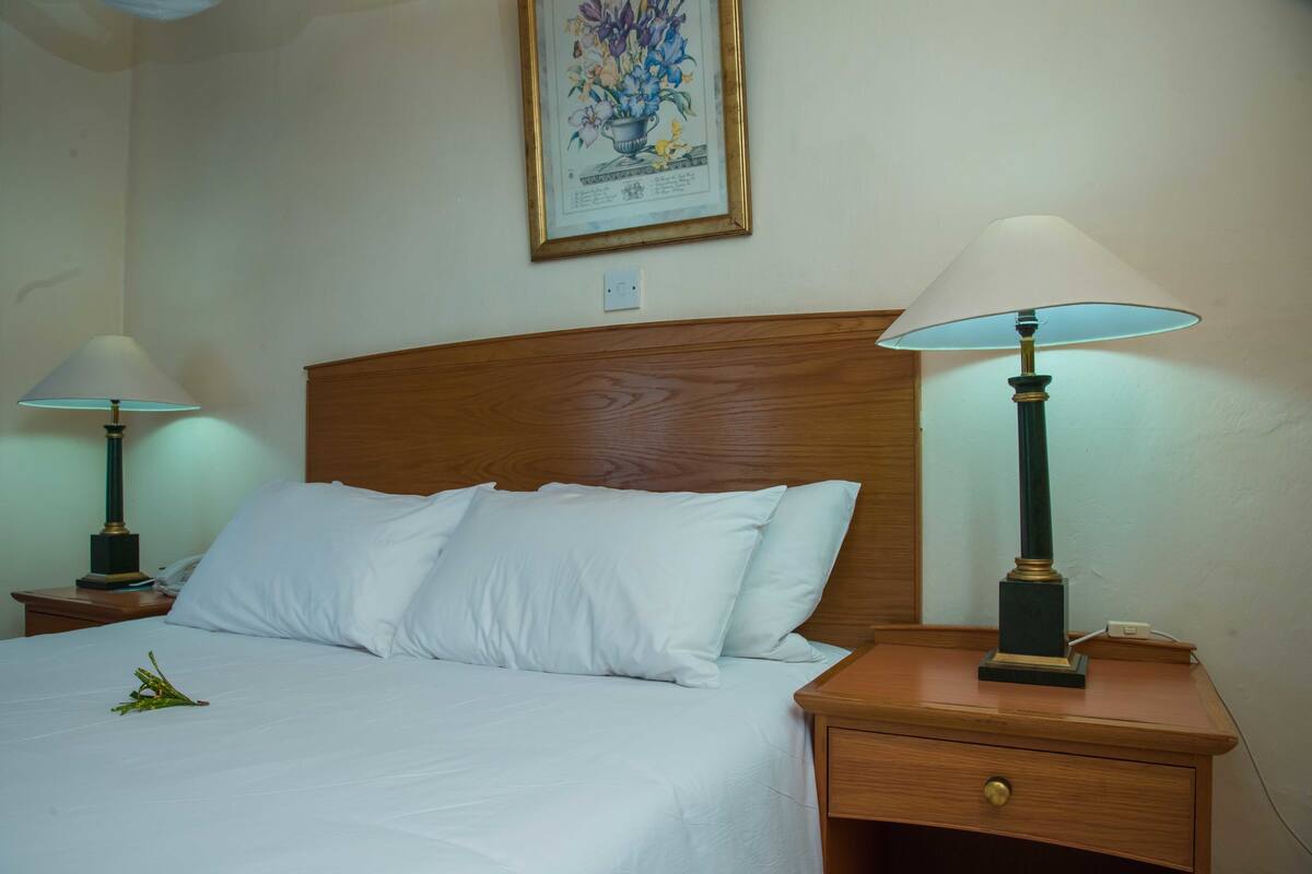 A neatly made bed with white linens, two bedside lamps, a wooden nightstand, and a framed floral artwork on the wall.
