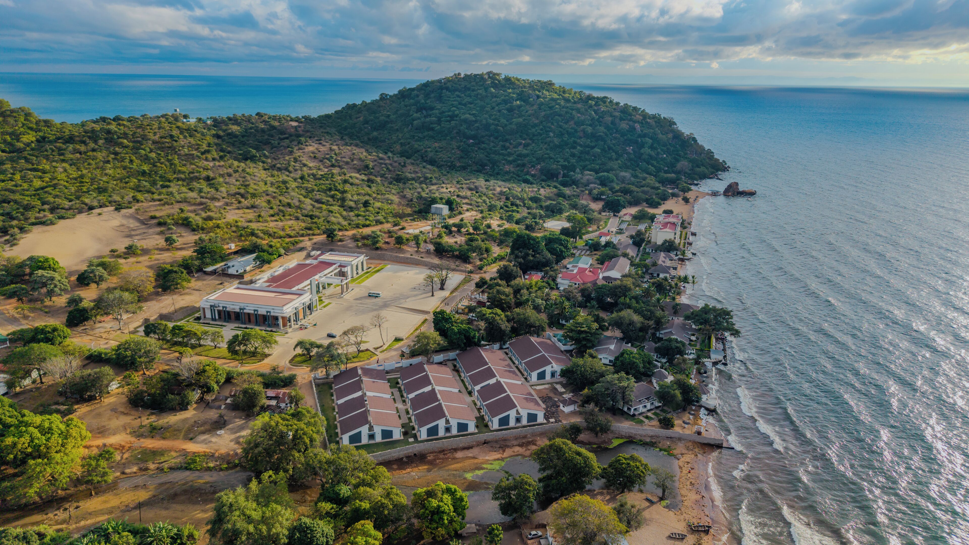 Vista aérea de una zona costera con un edificio grande, una playa y una colina boscosa.