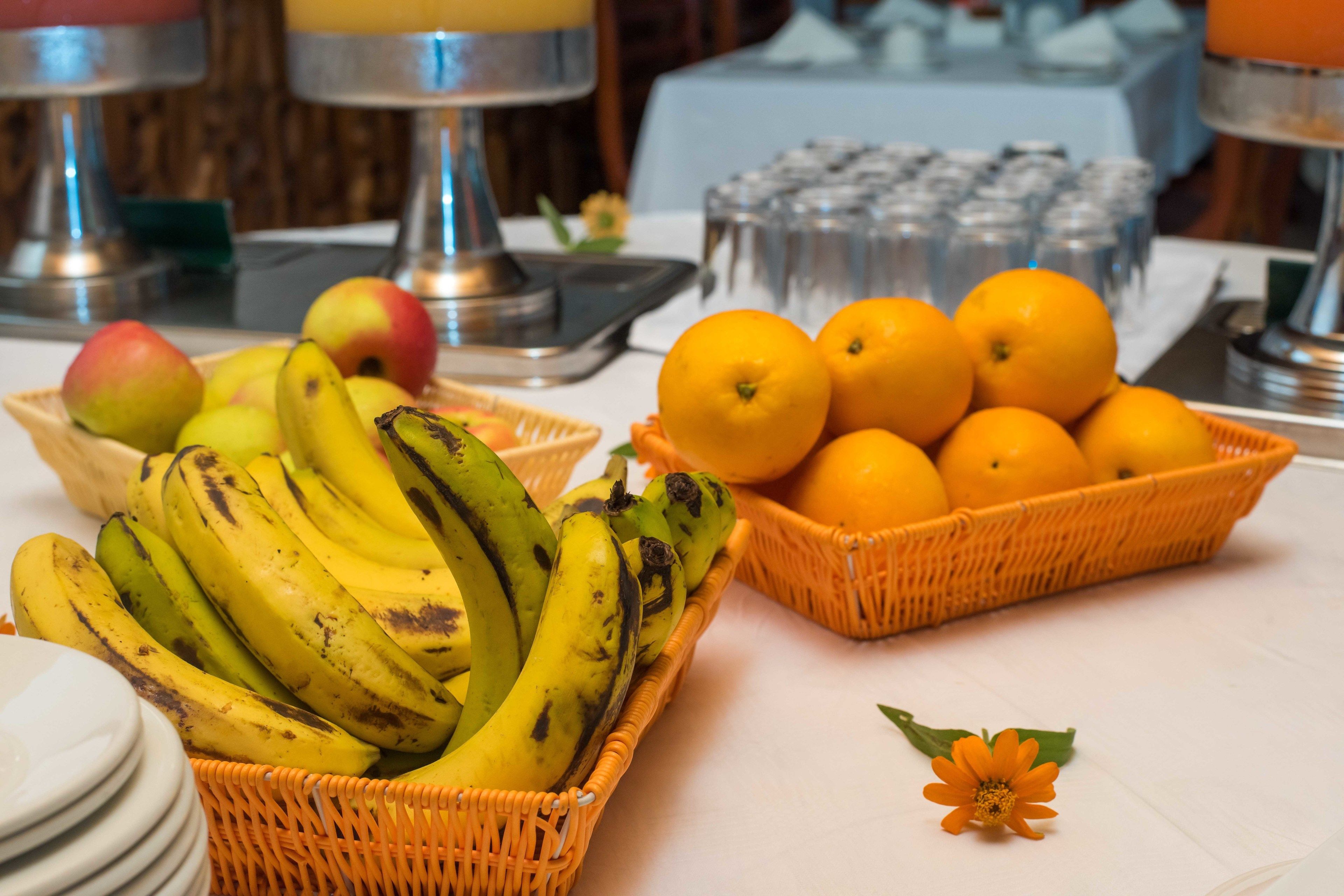 Una mesa con cestas de plátanos y naranjas, una pila de platos y una flor.