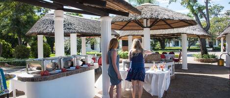 Un área de comedor al aire libre con mesas de buffet, cubierta por sombrillas de paja y columnas blancas.