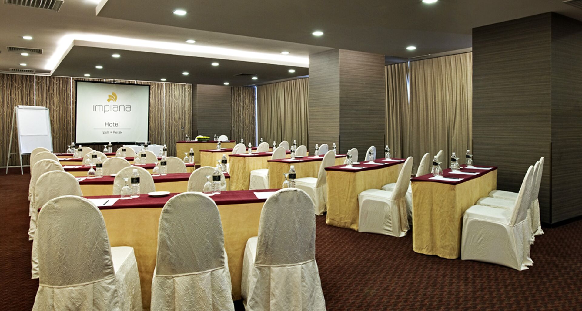 A conference room with round tables, white chairs, and a projection screen displaying "Implan Hotel".
