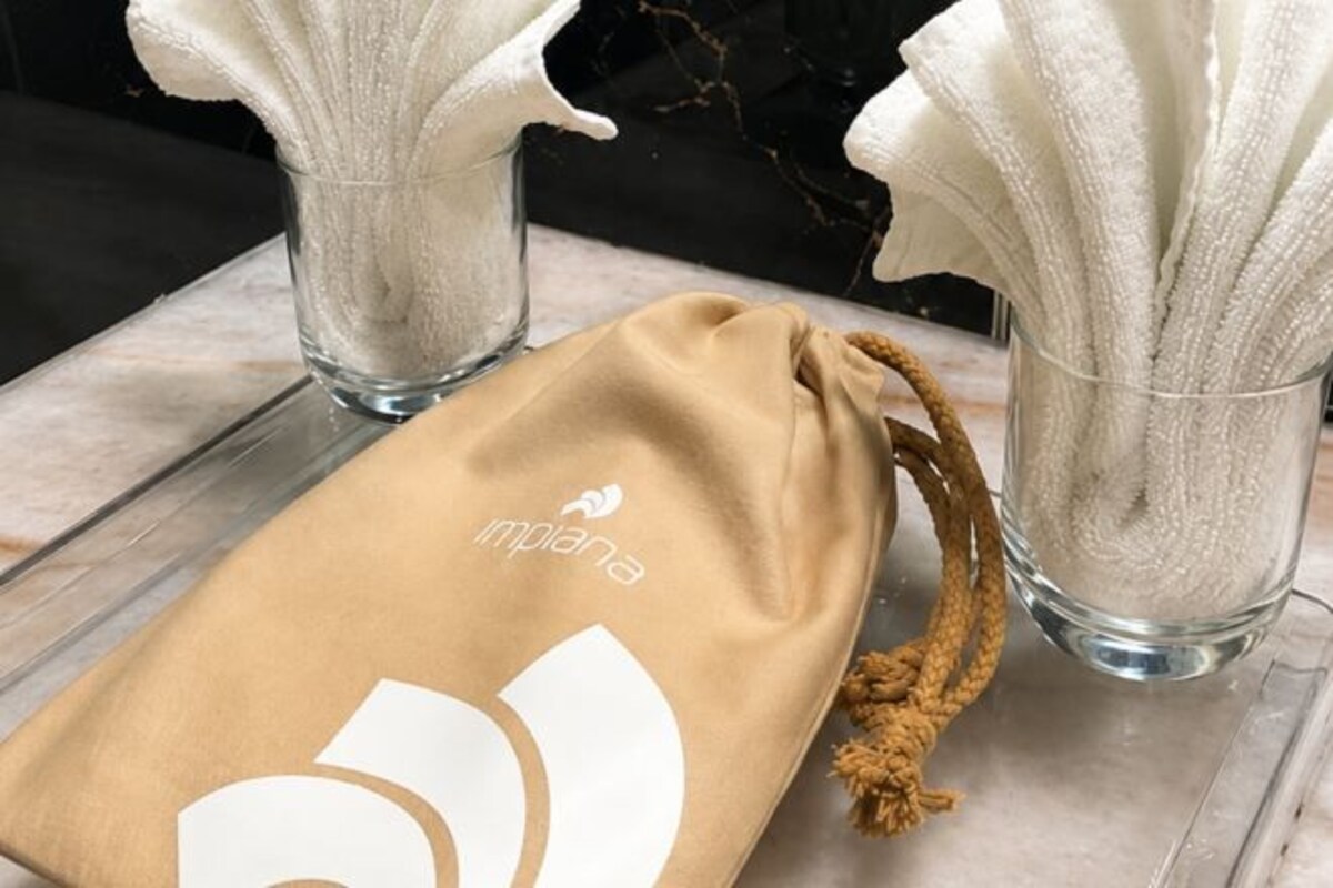 Una bolsa de tela marrón con cordón que lleva la palabra 'impana', sobre una superficie de mármol junto a dos jarrones de vidrio con servilletas blancas.