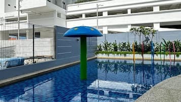 A modern building with a swimming pool, a blue umbrella, and a multi-story structure in the background.