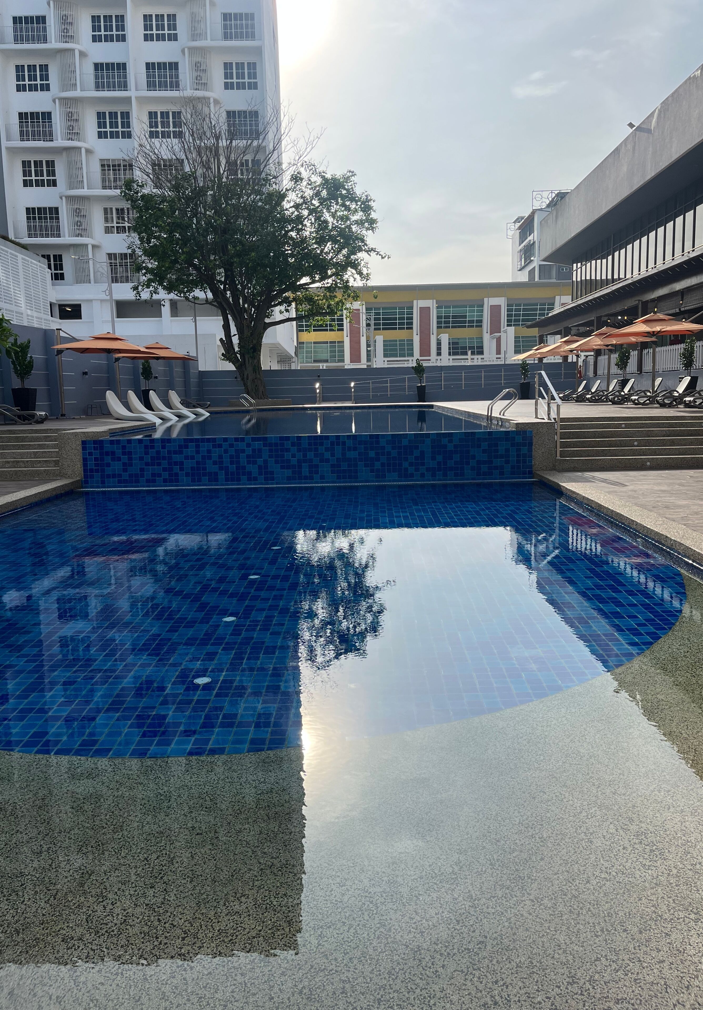 A swimming pool with blue tiles, surrounded by lounge chairs and umbrellas, with buildings in the background.