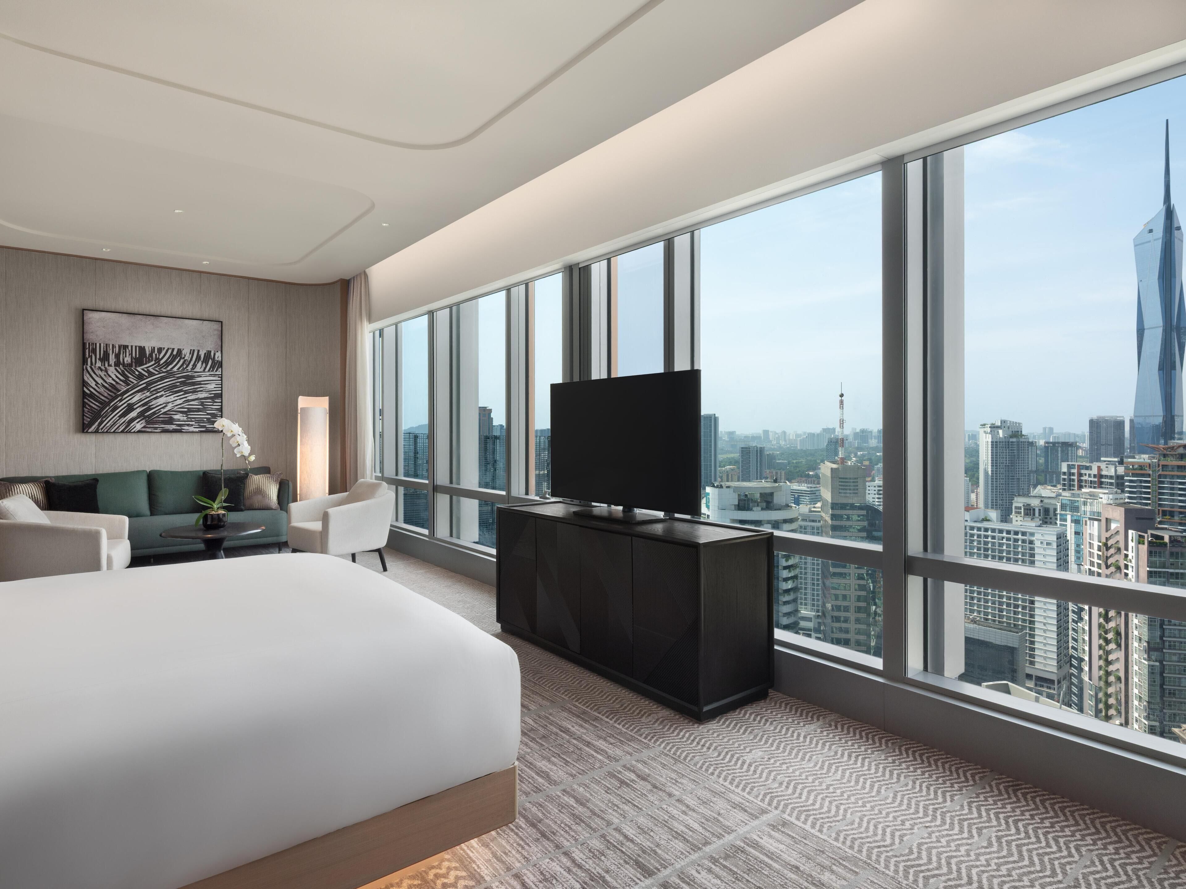 A modern hotel room with a large bed, a TV, a sofa, and a view of the cityscape.
