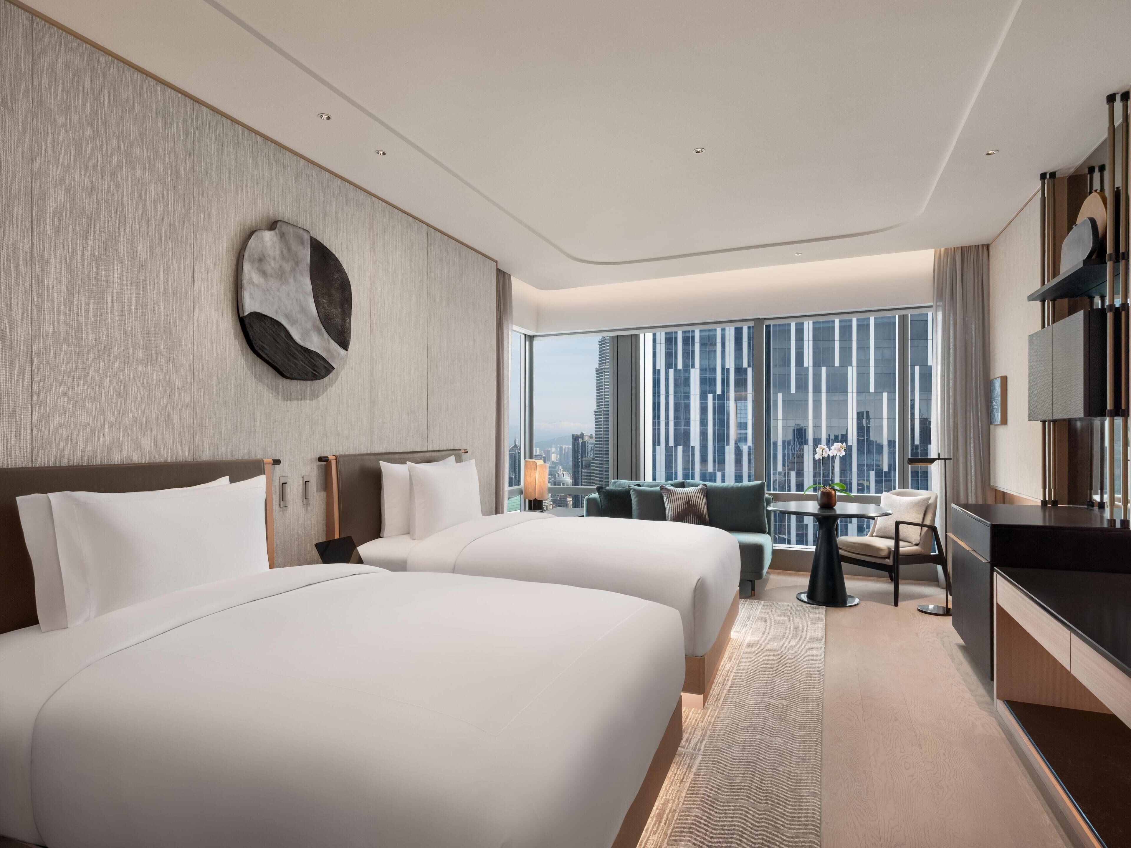 A hotel room with two beds, a desk, a chair, a TV, and a large window with a city view.