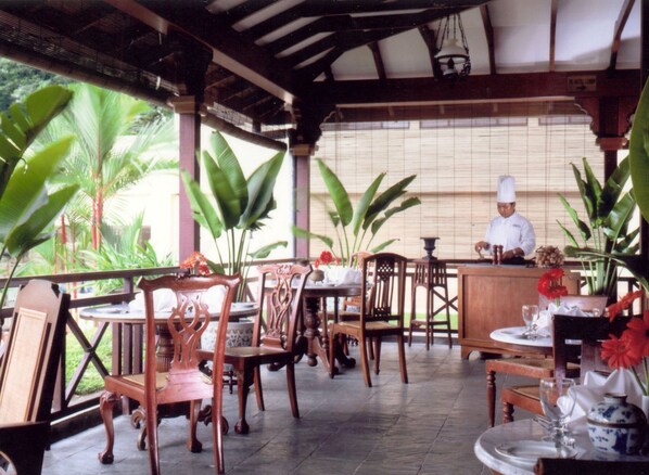 A restaurant with wooden chairs and tables, a chef at the counter, and large green plants. - Conrad Kuala Lumpur (Kuala Lumpur)
