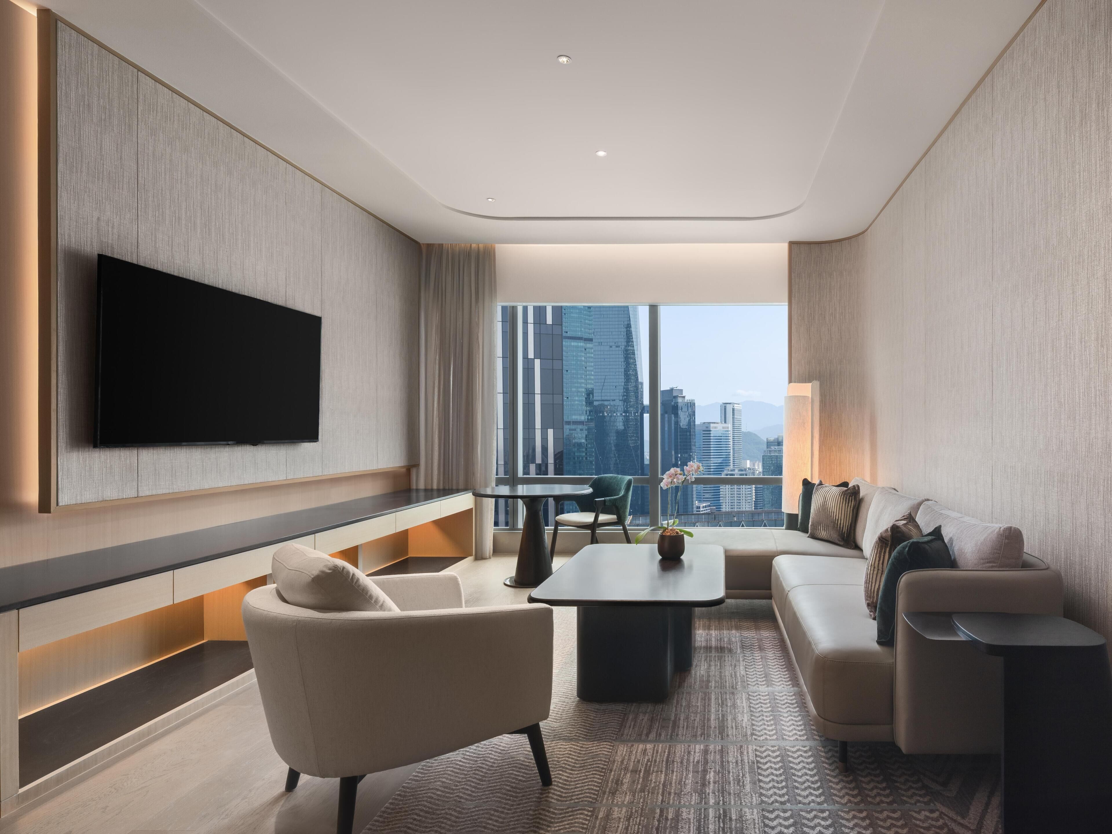 A modern hotel room with a flat-screen TV, a sofa, a coffee table, and a view of the cityscape.