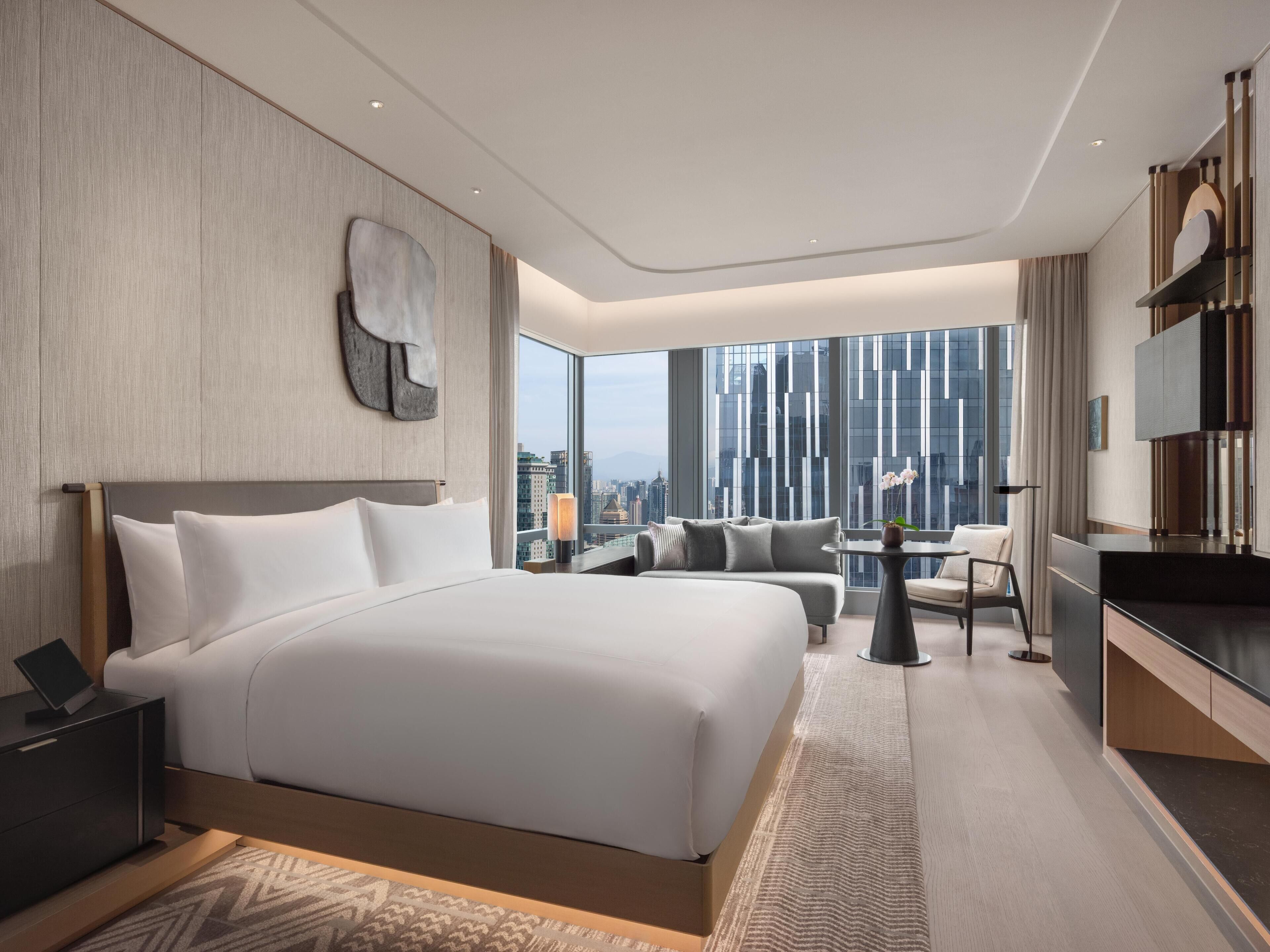 A modern hotel room with a large bed, a seating area, and a view of the cityscape.