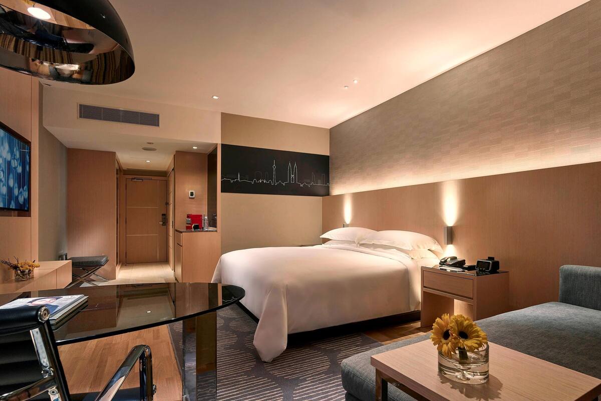 A modern hotel room with a bed, bedside table, lamp, and a glass table with a vase of flowers.