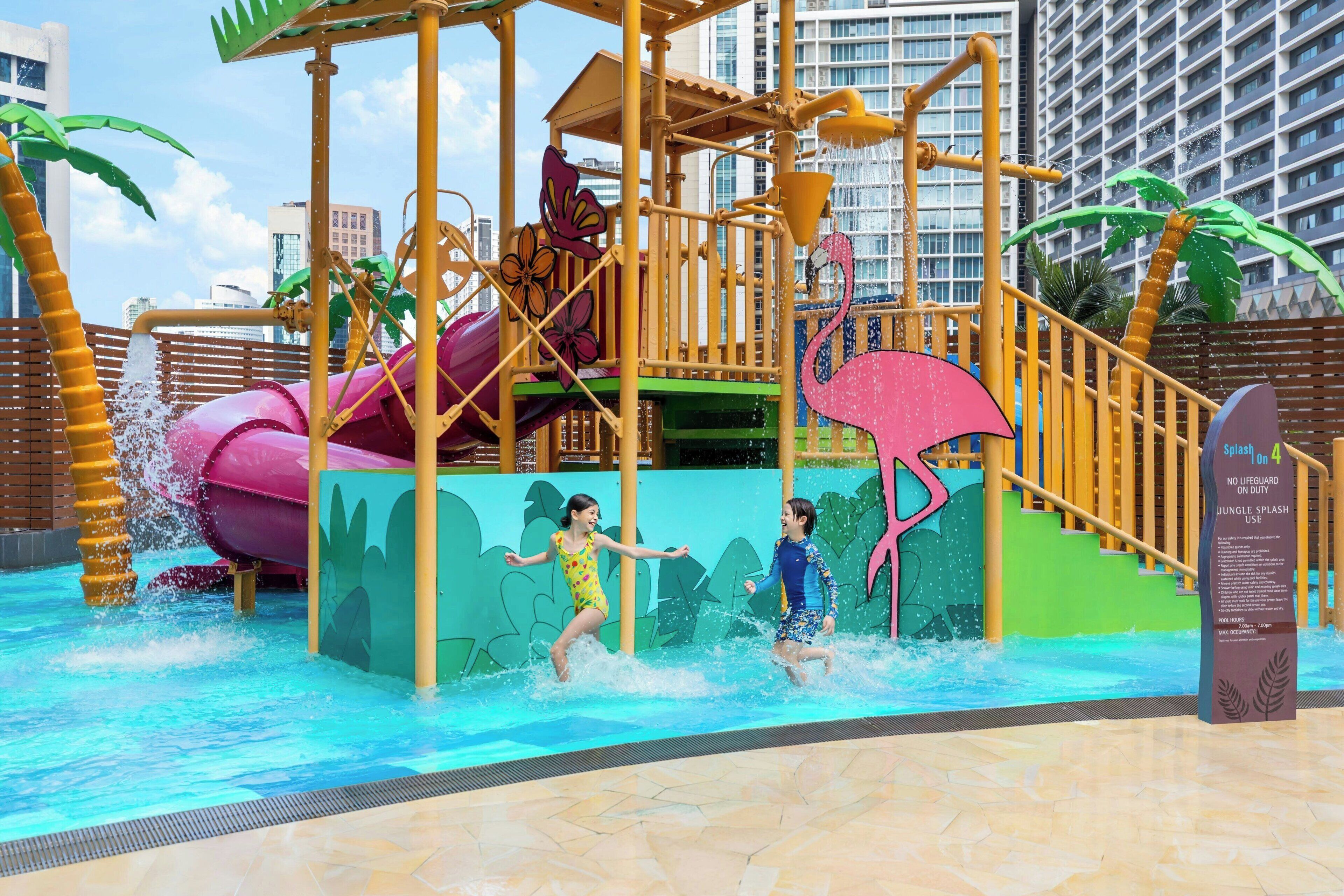 A water play area with slides and splash zones, surrounded by a wooden fence and palm trees.