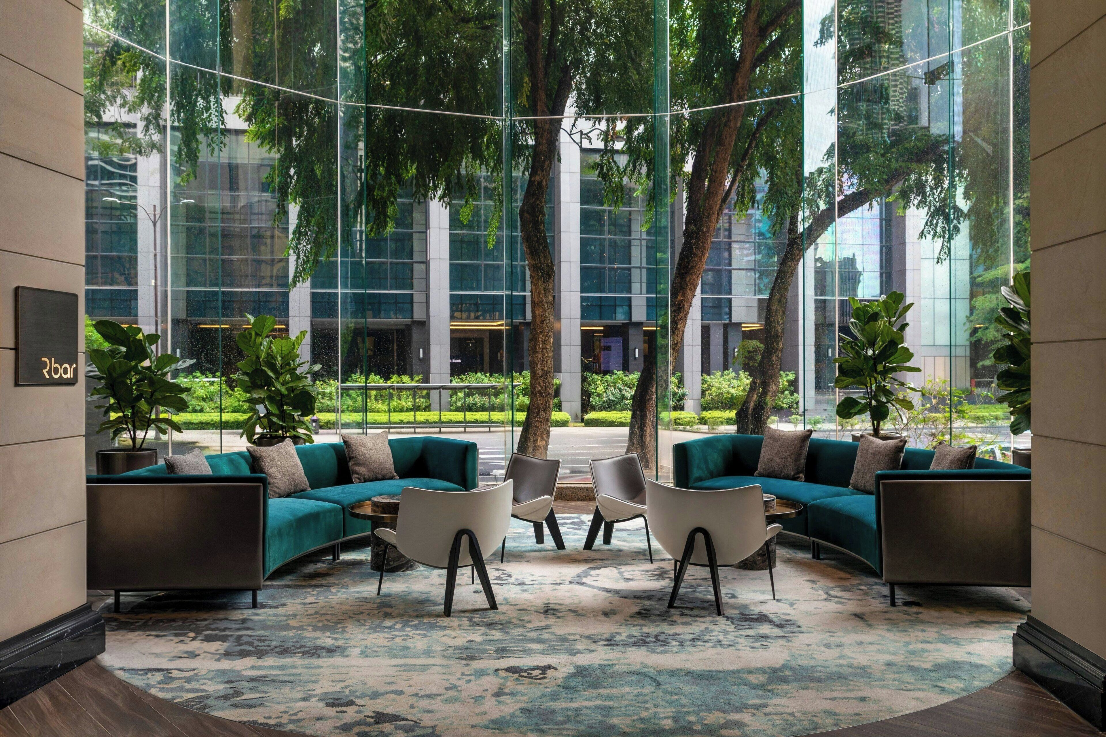 A modern lounge area with a curved sofa set, a coffee table, and chairs. There are potted plants and a large window offering a view of a building and trees.