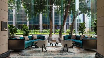 A modern lounge area with a curved sofa set, a coffee table, and chairs. There are potted plants and a large window offering a view of a building and trees.