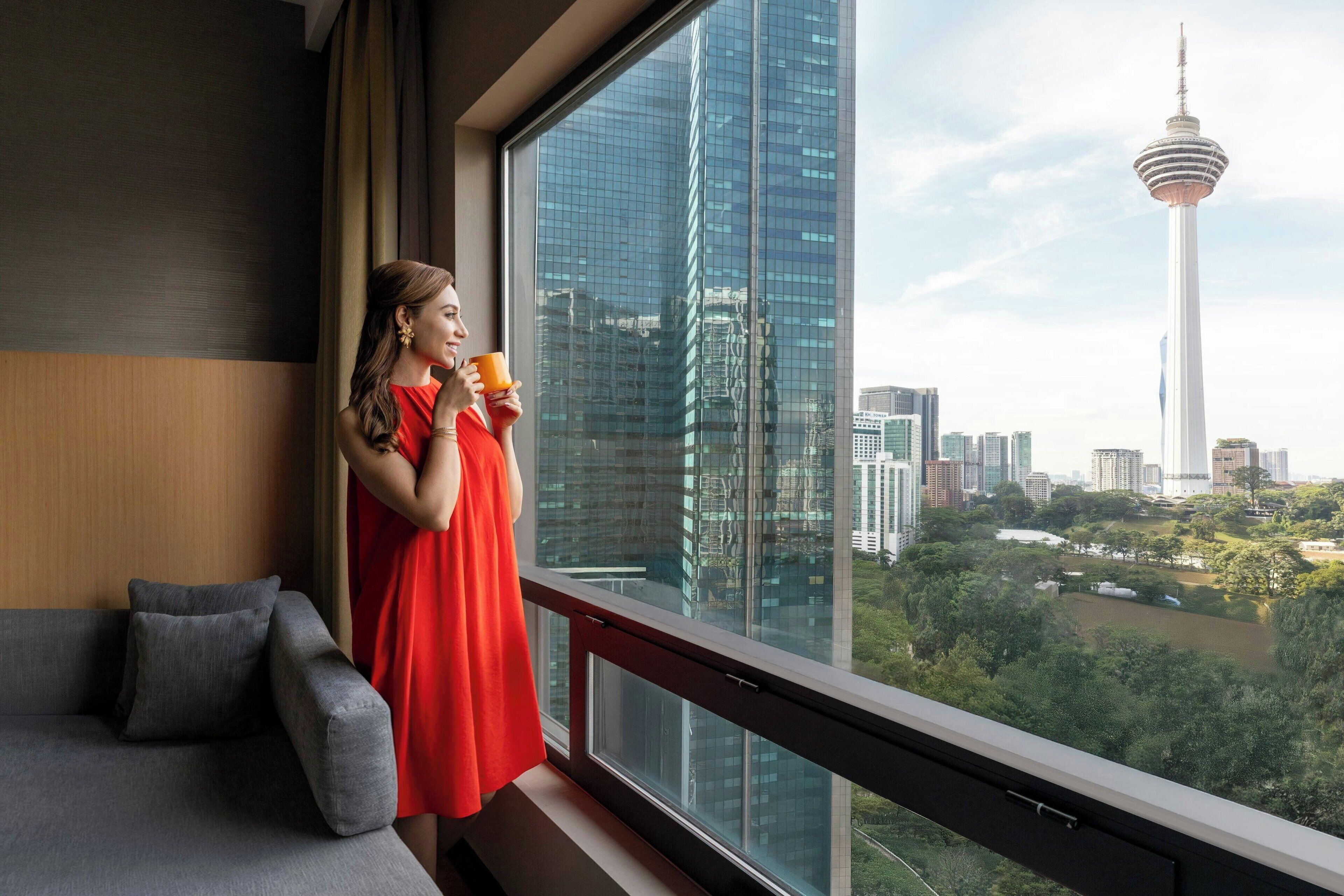 A person in a red dress standing by a window with a view of a cityscape and a tower.