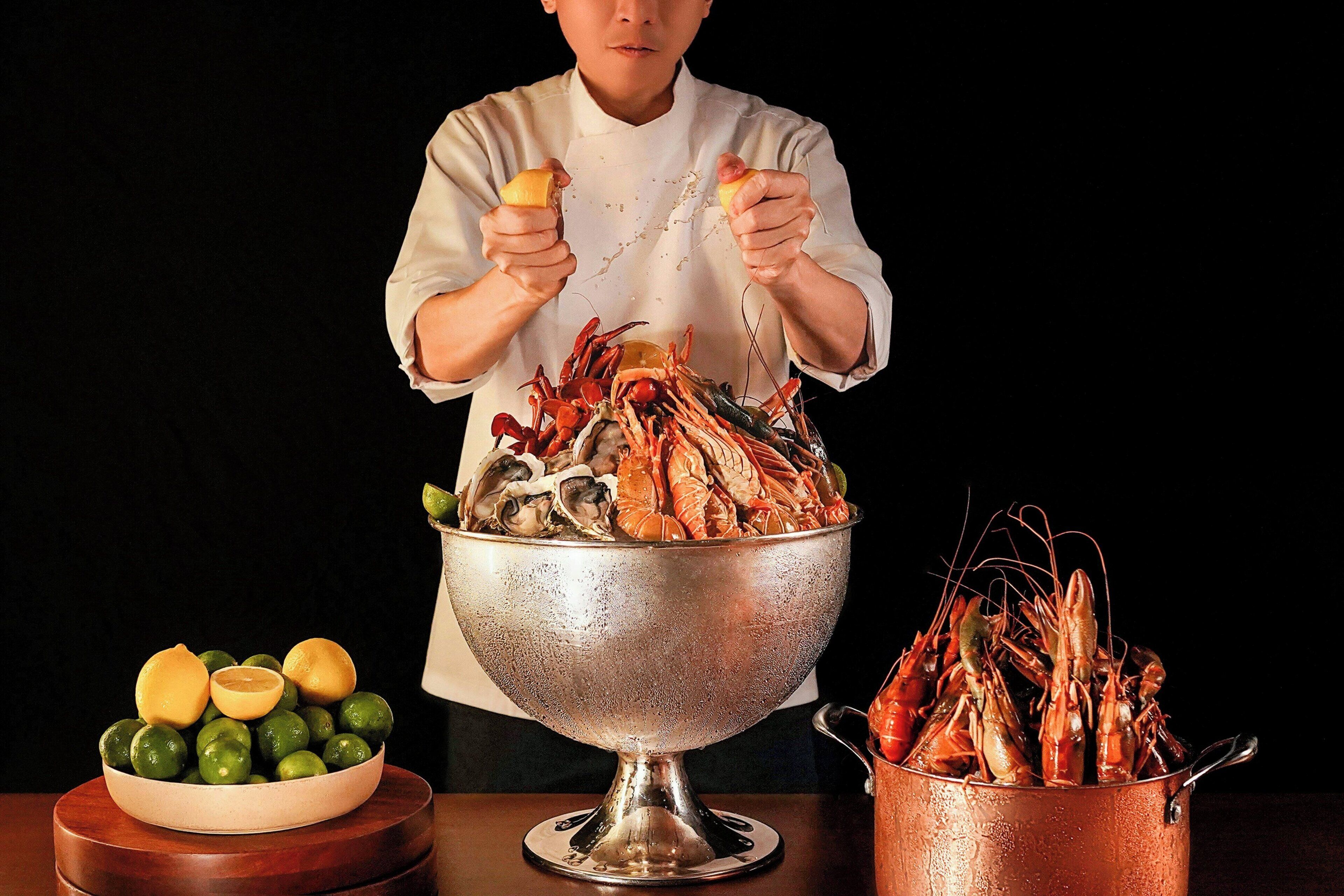 A chef holding a lobster and a lemon, with a bowl of green fruits and a pot of cooked lobsters.
