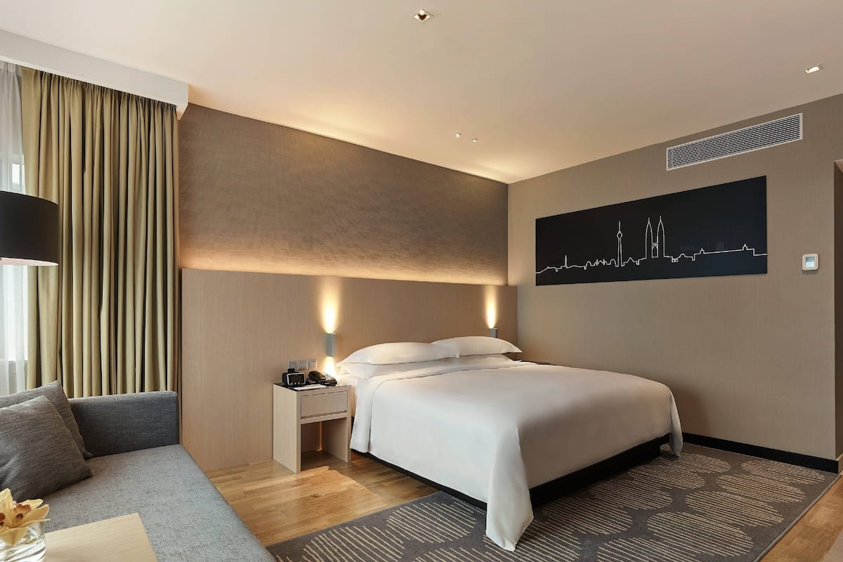 A hotel room with a large bed, a nightstand, a lamp, a sofa, and a cityscape painting on the wall.
