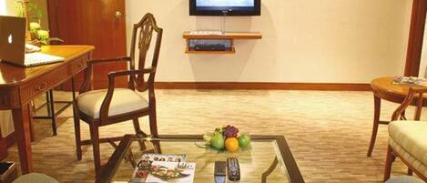 A hotel room with a wooden desk, a chair, a glass coffee table with magazines and a remote, a television on the wall, and a small shelf with a book.