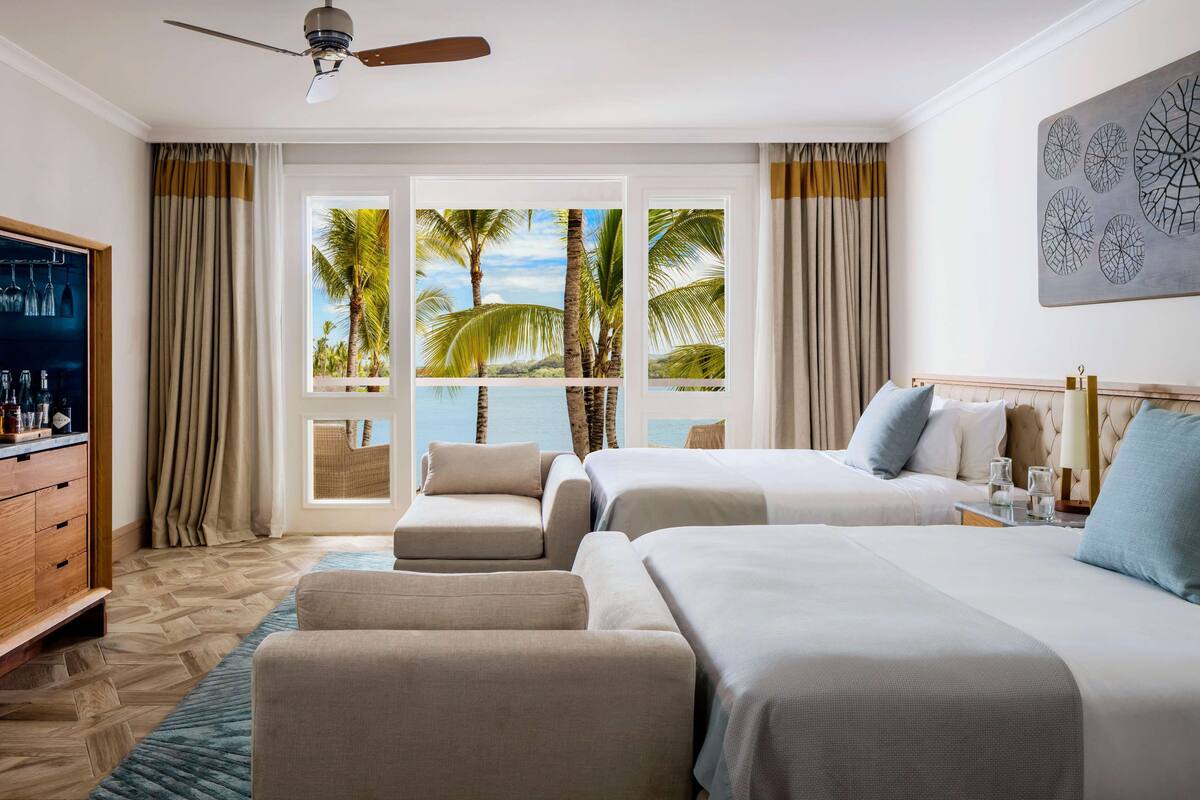 Habitación de hotel con dos camas, un sofá, un televisor y vistas a palmeras y al mar.
