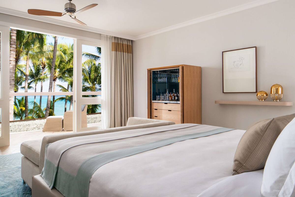 Habitación de hotel con una cama grande, zona de estar con vista a palmeras, minibar y ventilador de techo.