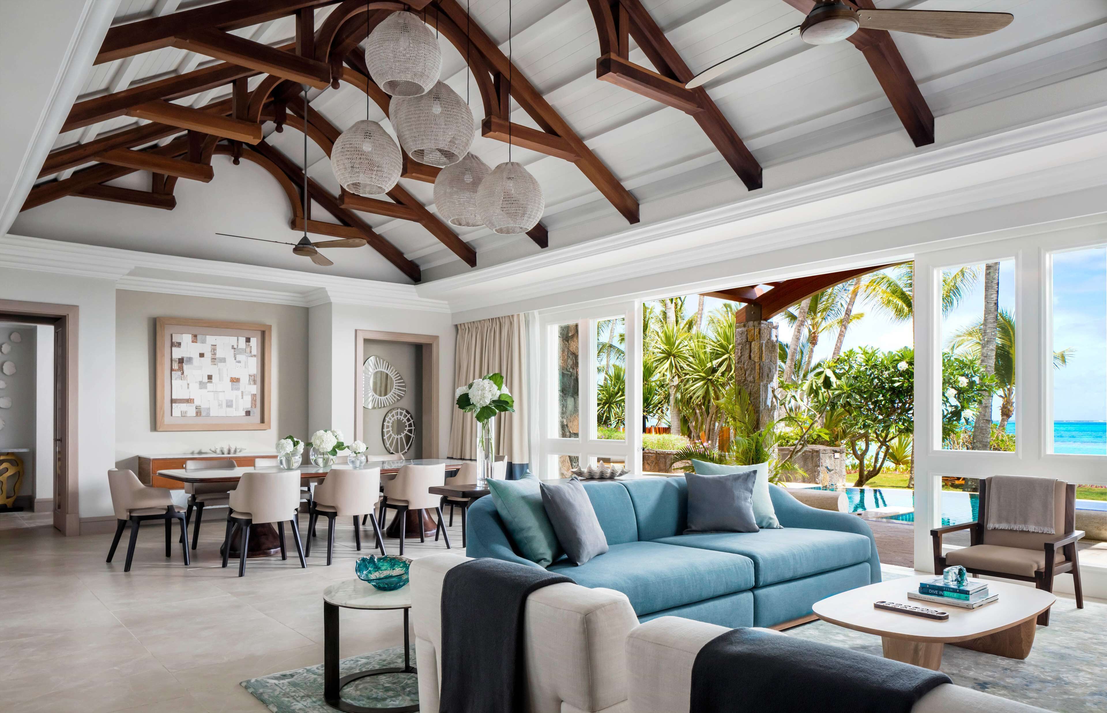 Amplia sala de estar con sofá azul, comedor y grandes ventanales con vista a un paisaje tropical.