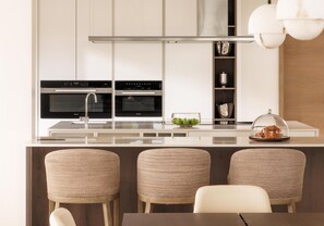 A modern kitchen with two ovens, a stainless steel range hood, and a central island with a glass top. There are three woven chairs and a white chair at the island.