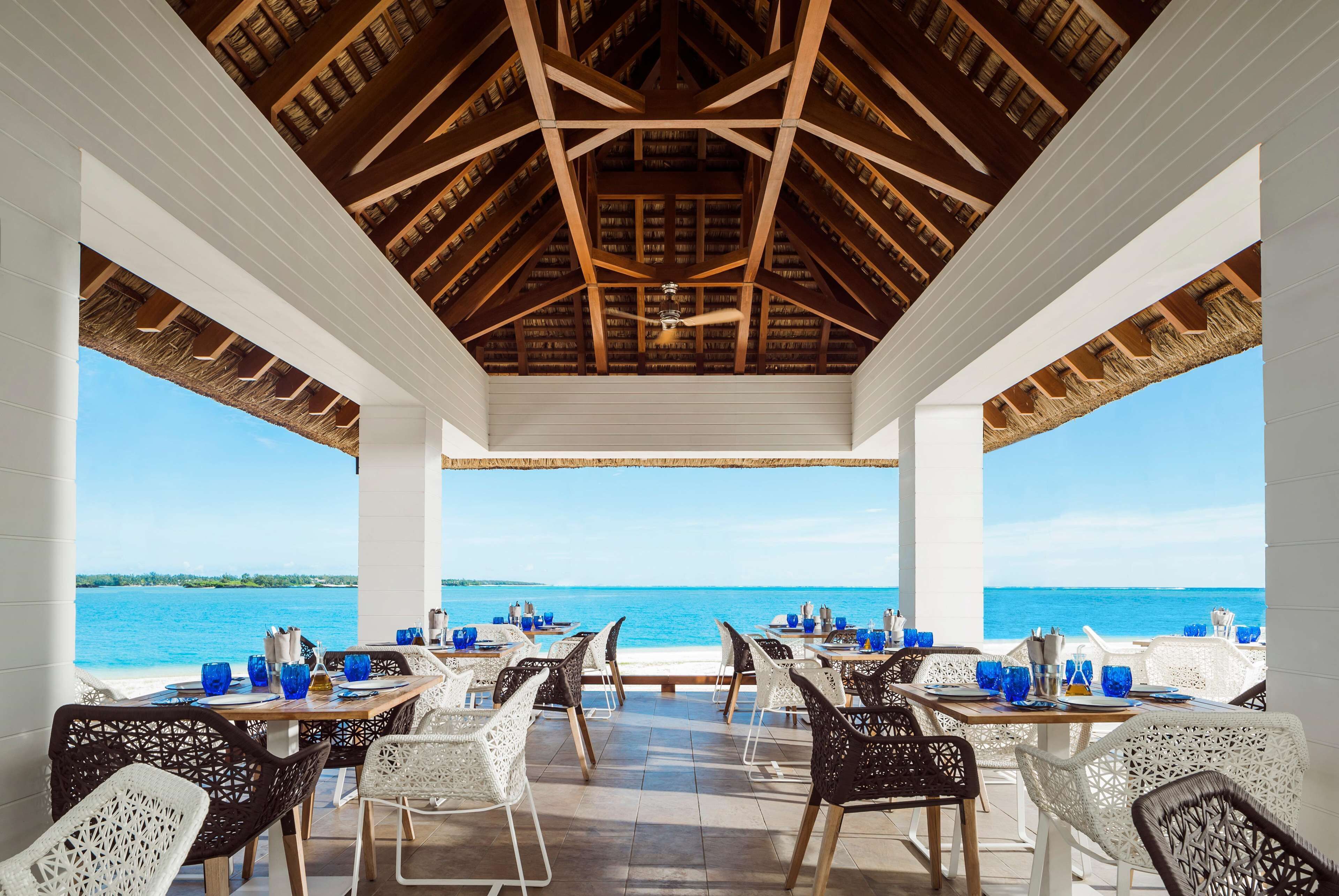 A covered outdoor dining area with tables and chairs, a ceiling with wooden beams, and a view of the sea.