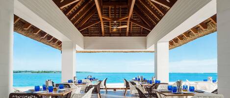 A covered outdoor dining area with tables and chairs, a ceiling with wooden beams, and a view of the sea.