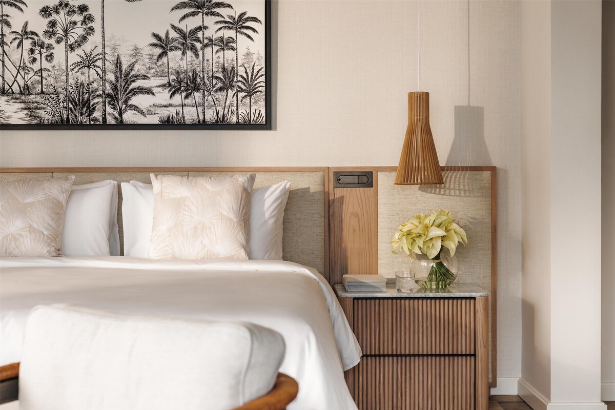 A neatly made bed with white linens, a wooden headboard, and a wall-mounted artwork depicting palm trees.