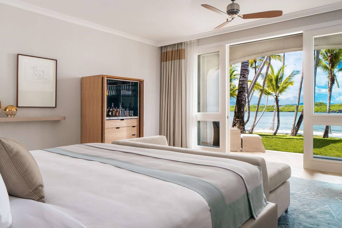 Un dormitorio con una cama grande, un minibar, un ventilador de techo y vistas a palmeras y al agua.