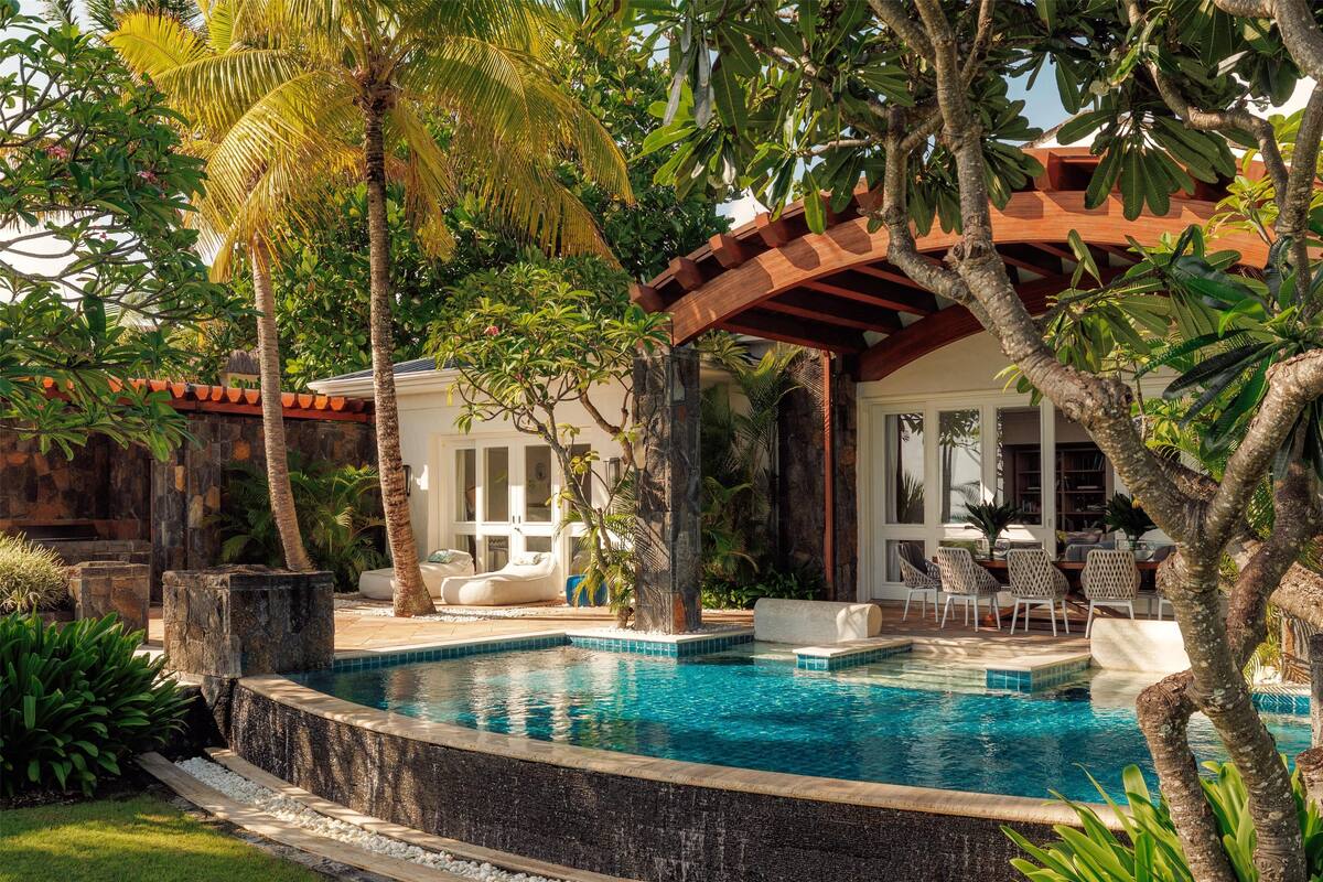 Un área de piscina con una pérgola de madera, palmeras y un muro de piedra.
