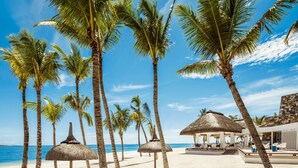 A beach with palm trees, a thatched-roof structure, and a building with outdoor seating. - One&Only Le Saint Géran (Belle Mare)