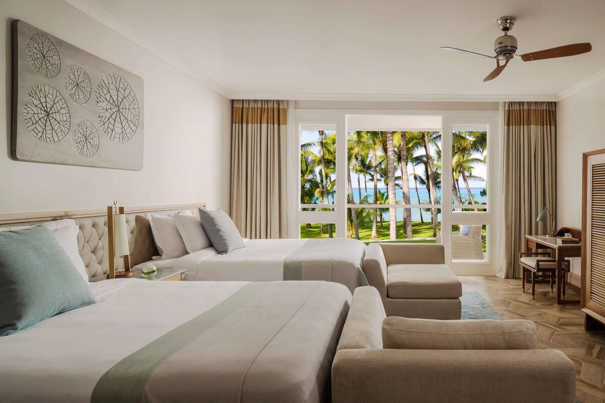 Habitación de hotel con dos camas, un escritorio y vistas a palmeras y al océano.
