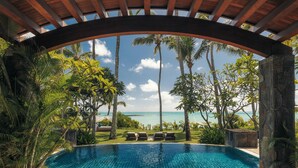 A pool with lounge chairs under a wooden pergola, surrounded by palm trees and a view of the ocean. - One&Only Le Saint Géran (Belle Mare)