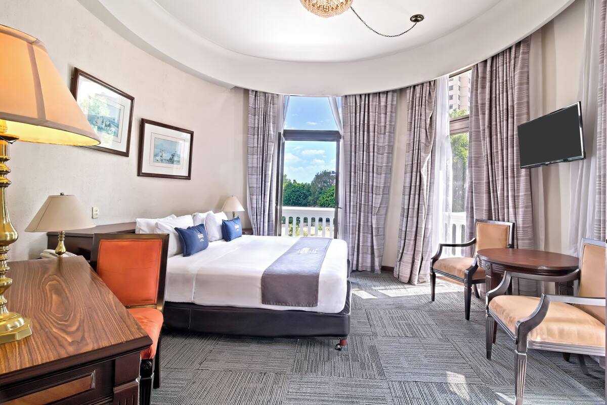 Habitación de hotel con cama, escritorio, silla, televisión y ventana con cortinas.