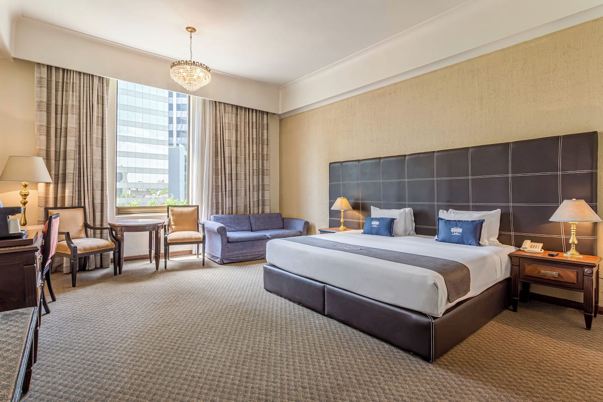 Habitación de hotel con una cama grande, un escritorio con lámpara, un sofá y vistas de la ciudad a través de amplios ventanales.