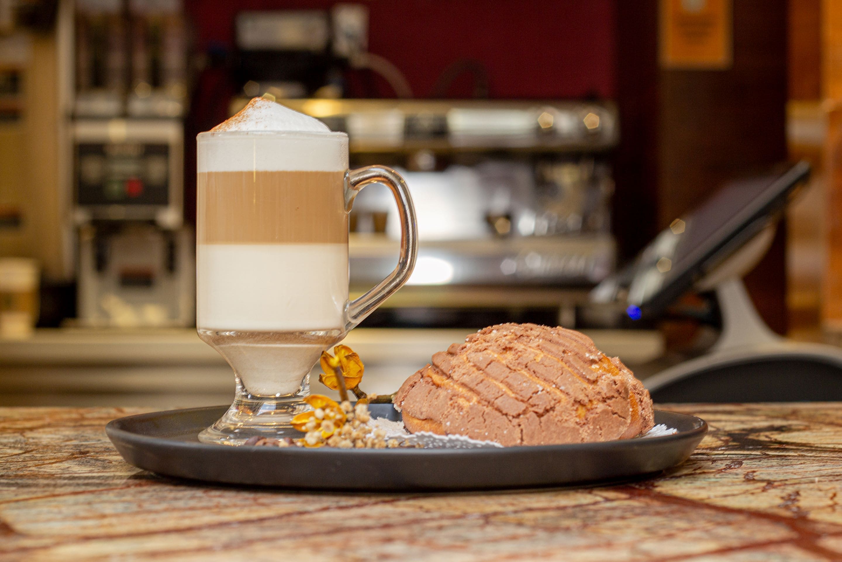 A dessert consisting of a layered beverage and a pastry on a plate, with a blurred background of a coffee machine and counter.