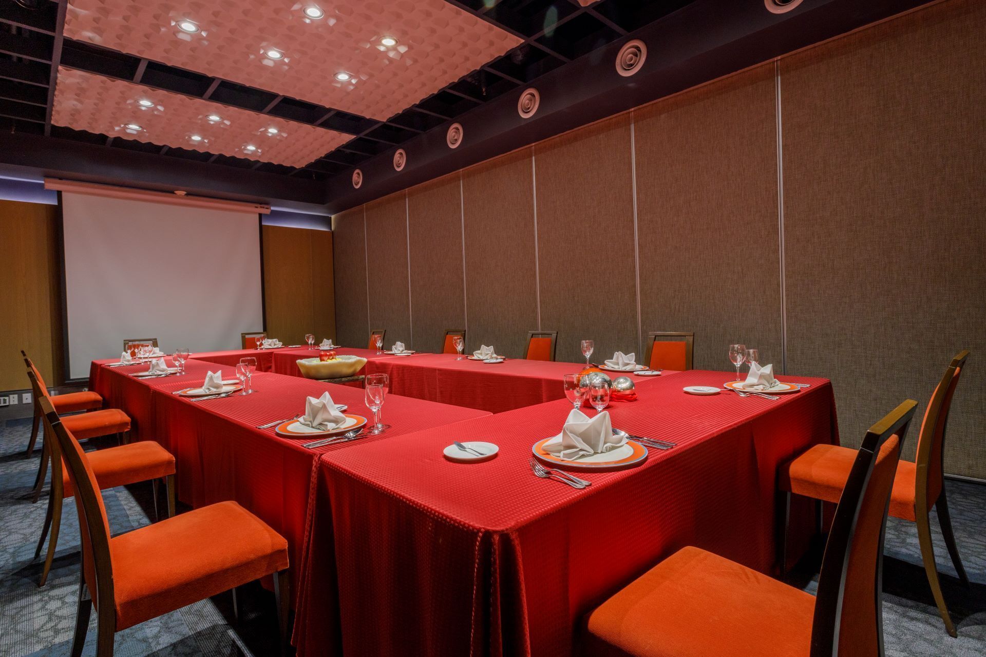 Sala de conferências com mesas vermelhas e cadeiras laranja, equipada com toalhas de mesa brancas e talheres.