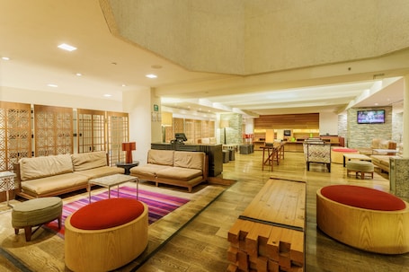 A spacious lobby with a sofa set, round wooden ottomans, a reception desk, and a TV mounted on the wall.. Camino Real Aeropuerto Mexico