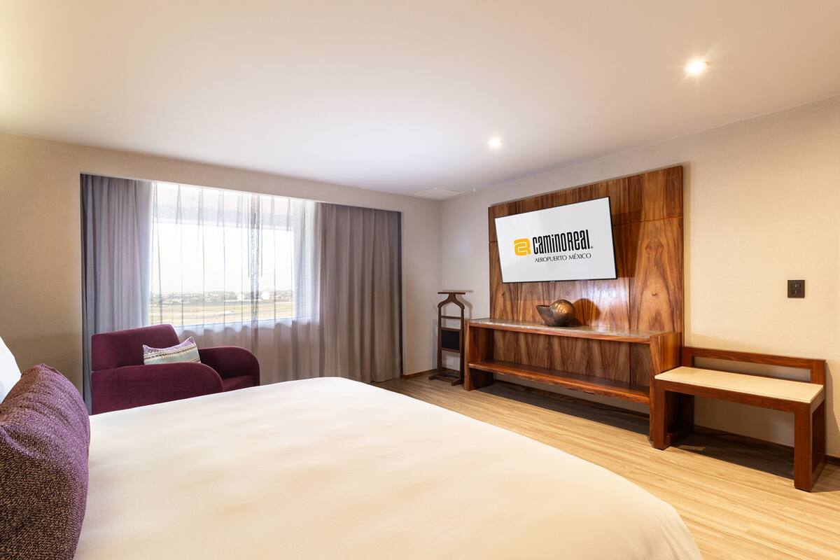A hotel room with a large bed, a seating area with a bench, a wooden shelf, and a large window with curtains.