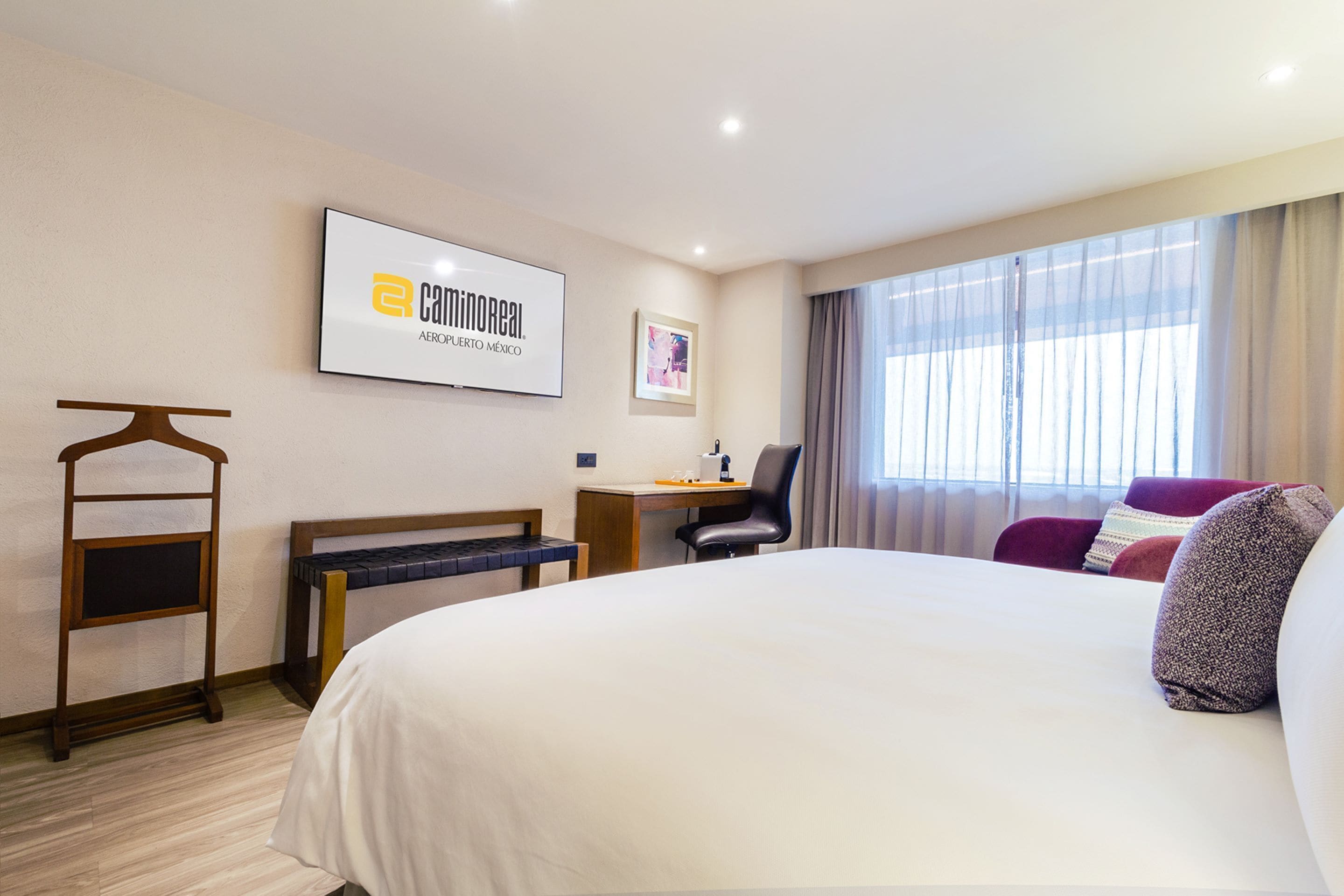 Habitación de hotel con una cama grande, un escritorio con silla, un televisor montado en la pared y un ventanal con cortinas.